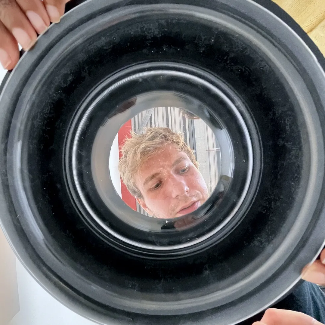 A distorted circular reflection seen through a black bowl, with a person’s face appearing in the center, surrounded by concentric rings of light and shadow. Hands hold the bowl at the edges.