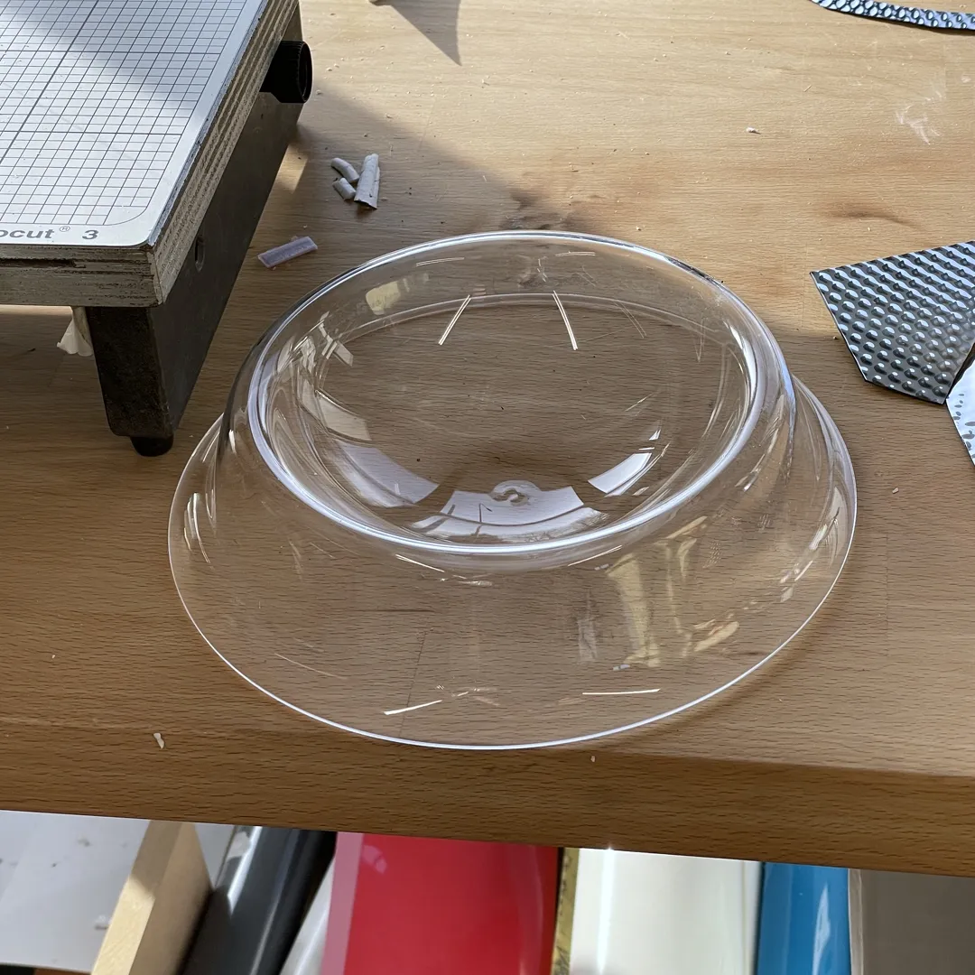 Transparent plastic bowl resting on a wooden table, surrounded by crafting materials, including paper and textured surfaces.